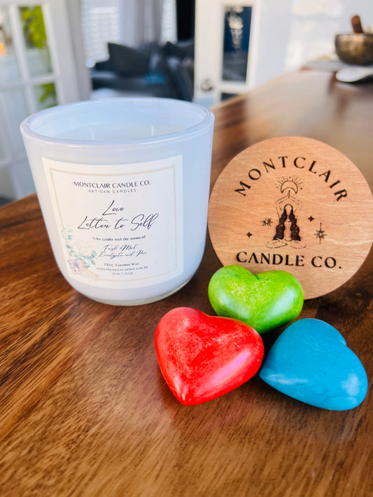 A white candle labeled 'Letter to Self' from Montclair Candle Co. is displayed next to colorful heart-shaped wax decorations on a wooden surface.